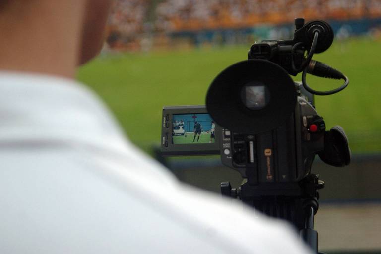 Fotografia colorida mostra, em primeiro plano e desfocado, o ombro de um cinegrafista que regista uma partida de futebol. Em segundo, há sua câmera e a imagem do campo. Ao fundo, o gramado verde