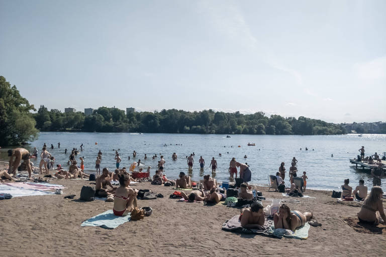 Pessoas tomam sol, sentam na areia e entram na água em um lago.