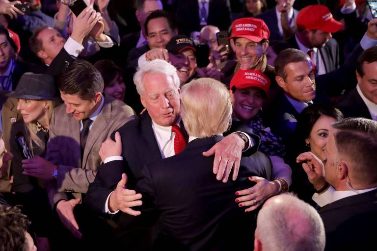 Em meio a multidão, homem grisalho, frente à câmera, abraça Donald Trump, visto de costas