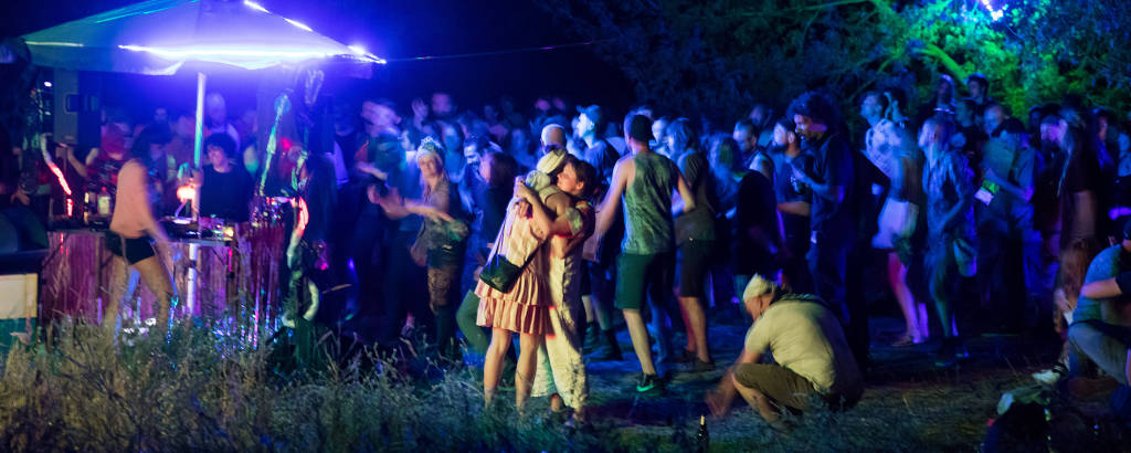 Jovens aglomerados em festa se abraçam. Luzes de neon azuis iluminam local, com árvores