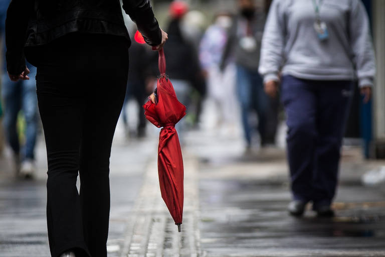 Mulher anda em calçada segurando um guarda-chuva fechado. Um homem vem em sentido contrário