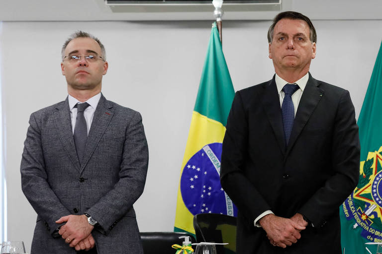 André Mendonça e Jair Bolsonaro estão em pé diante de uma mesa; ao fundo, uma bandeira do Brasil hasteada