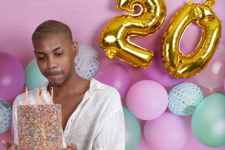 Um homem segura um bolo colorido, com uma parede rosa ao fundo e balões com numero 20