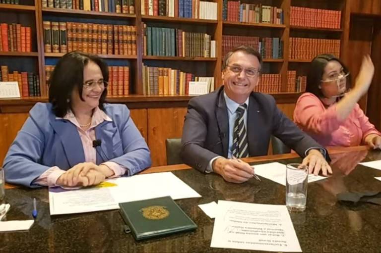 Em frente a uma estante de livros, a ministra Damares Alves, à esquerda, o presidente Jair Bolsonaro, no centro e a tradutora de Libras se sentam a uma mesa. A ministra e o presidente riem.