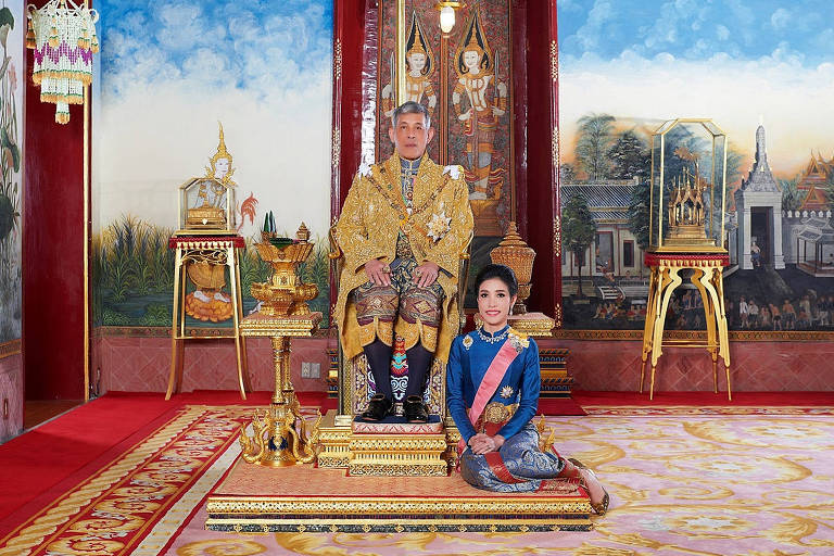 Sentado em um trono, o rei da tailândia posa ao lado da consorte real, sentada aos seus pés, no chão.