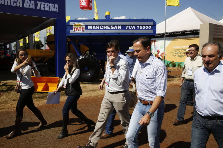 três homens de calça e camisa andam em rua de feira agrícola