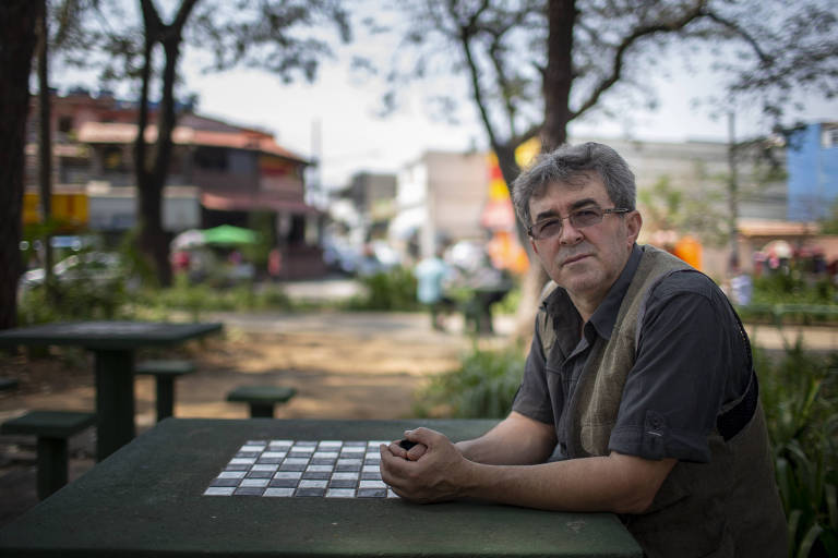 Senhor de óculos e cabelos brancos com os braços sobre a mesinha de uma praça, que tem no meio um tabuleiro de xadrez