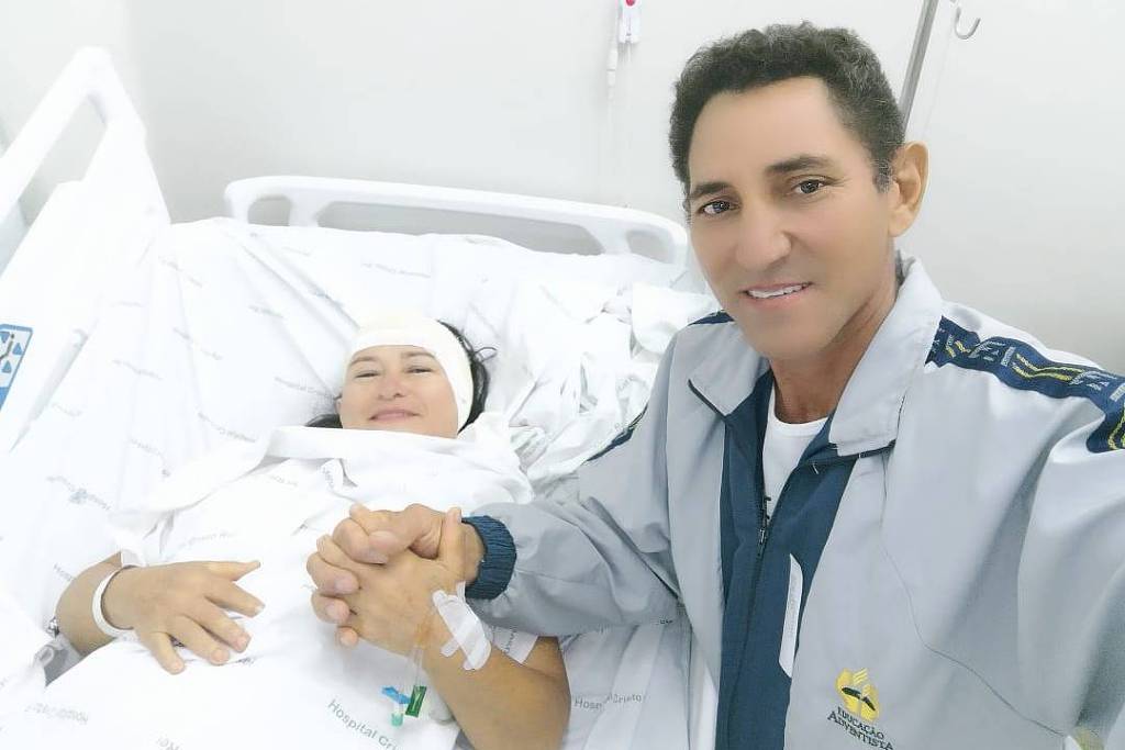 Mulher com faixa rodeando a cabeça acima dos olhos, deitada em cama de hospital, ela sorri e está de mão dadas com homem de pé que também sorri