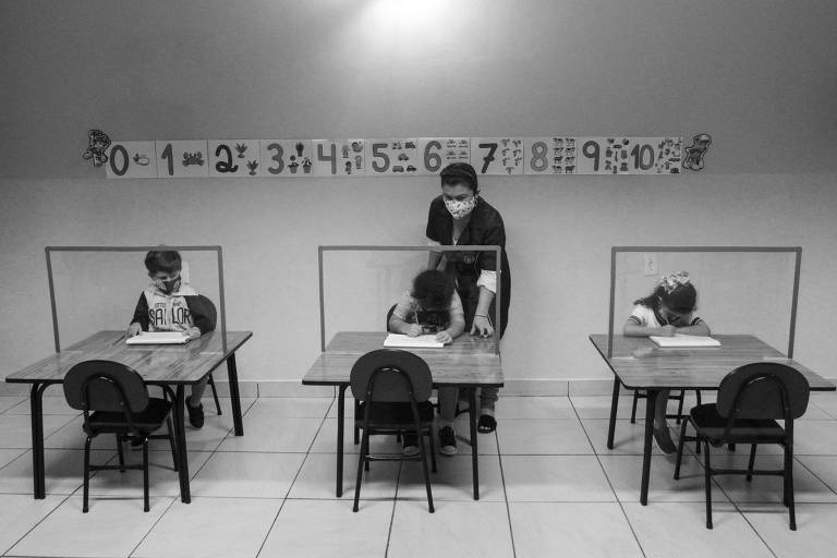Três crianças estudando na classe com máscaras de proteção em mesas afastadas, acompanhadas por um professor