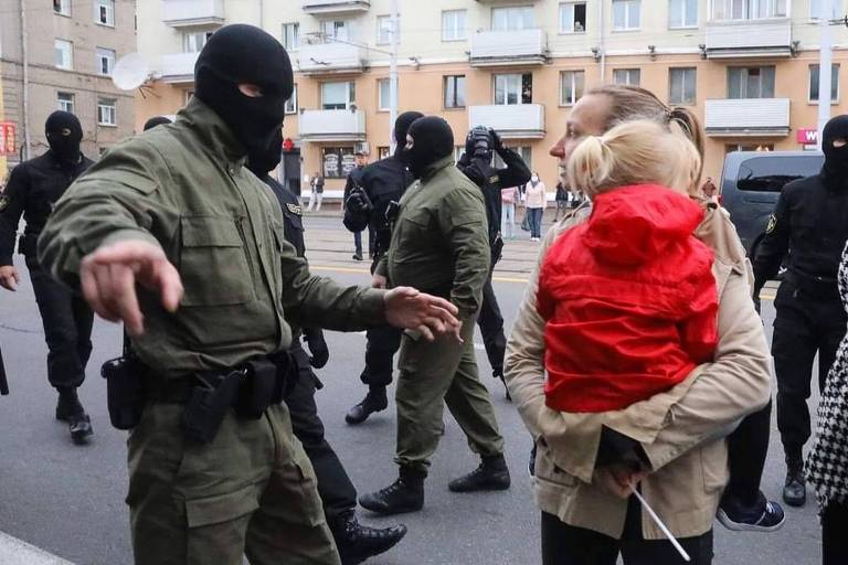Policial mascarado gesticula mandando mulher de rabo de cavalo com criança de casaco vermelho no colo sair da rua