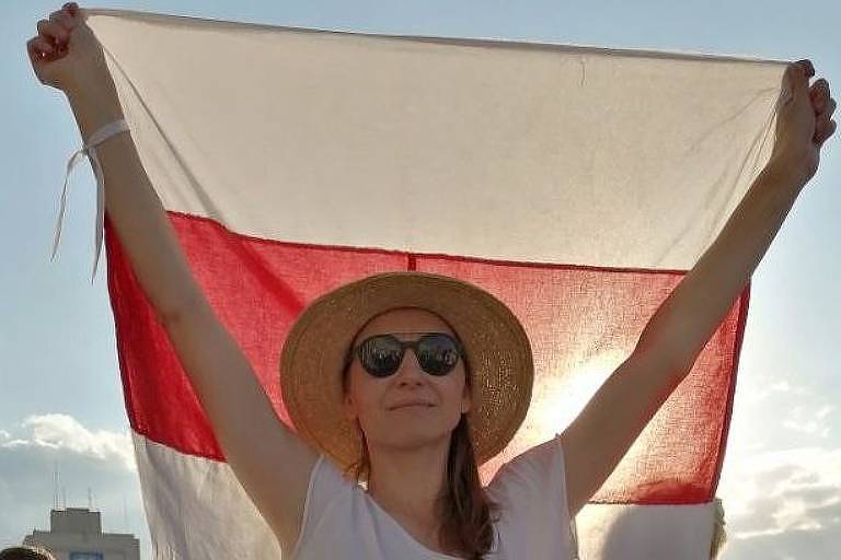 Mulher de chapeu e óculos escuros levanta com os dois braços bandeira branca, vermelha e branca