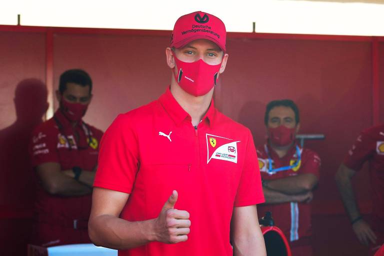 Mick Schumacher, de máscara, camisa e bonés vermelhos, acena para foto
