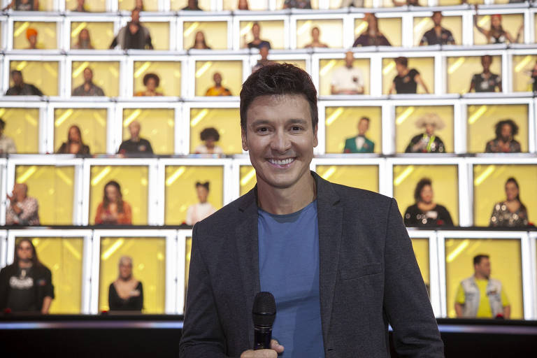 Faro veste paletó escuro e camiseta azul; ele segura microfone com a mão direita; ao fundo, painel com imagens de várias pessoas em retângulos de fundo amarelo