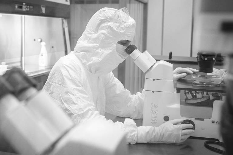 Cientista com trajes brancos olha em microscópio dentro de laboratório 