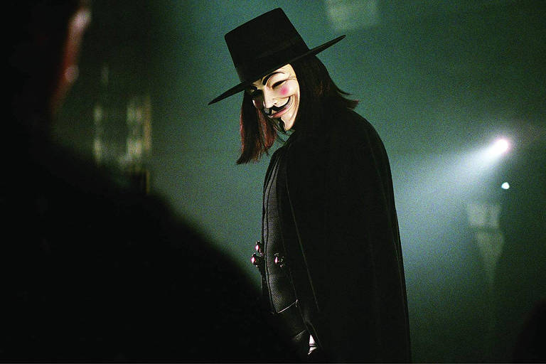 Um homem com máscara branca sorridente, cabelos longos, roupa preta com capa e chapéu encara a câmera em um cenário escuro levemente iluminado por uma lanterna. 