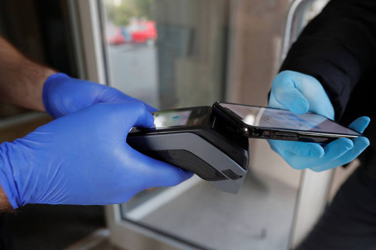 Mãos com luva azul seguram máquina de cartão, enquanto outra pessoa, também de luva azul (mais clara) segura celular para fazer pagamento por aproximação