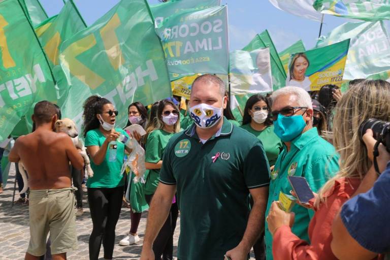 Homem ao centro de máscara e camiseta verde escuro com banddeiras verdes de campanha atrás dele e apoiadores