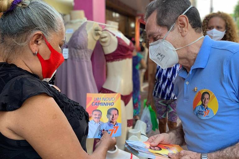 Um homem à direita de máscara e camisa azul conversa com uma mulher de máscara. Os dois mostram santinhos de campanha eleitoral.