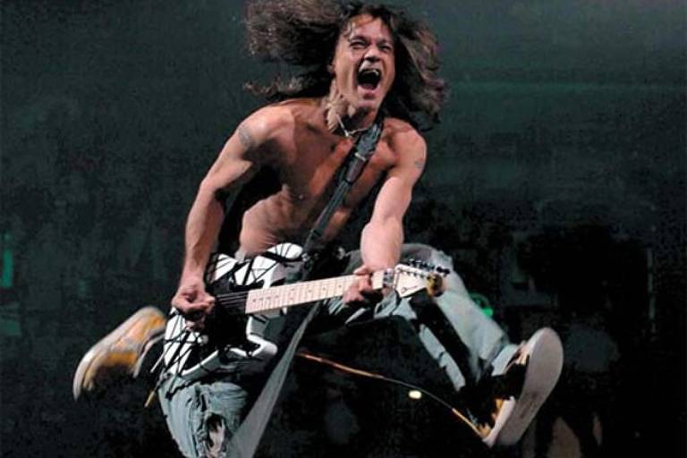 Eddie Van Halen, sem camisa e tocando sua guitarra, dá um salto e grita em show