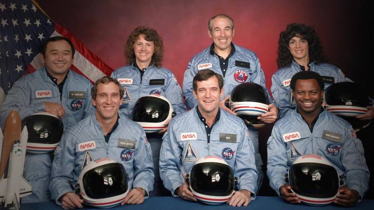 Duas mulheres e cinco homens posam para foto, com roupas de astronauta e capacete, e a bandeira dos EUA ao fundo