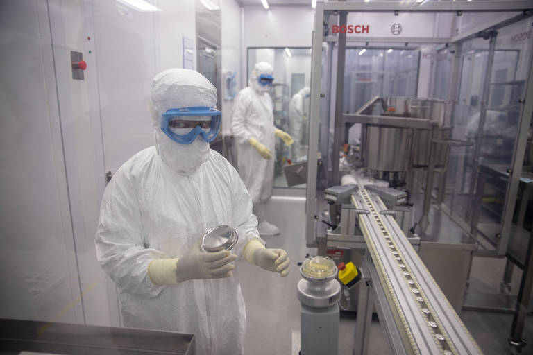 Cientista de uniforme de proteção branco, com óculos, segura pote em laboratório; ao fundo, outra pessoa com a mesma vestimenta