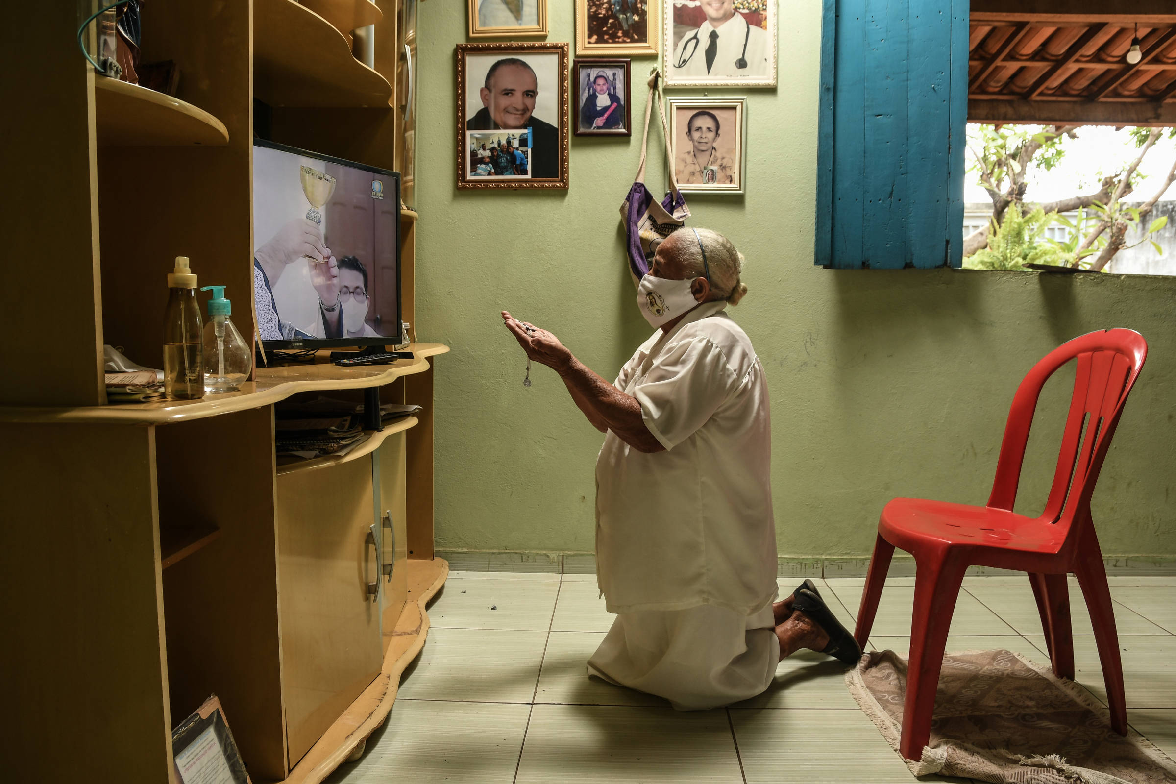 Com máscara, a aposentada Joana Chagas, 80, ajoelhada em sala de casa assiste à missa pela televisão em Juazeiro do Norte (CE) por causa da pandemia de Covid-19