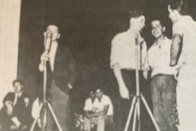 À esq. uma pessoa cantando de pé em um microfone com pedestal; à direita, três pessoas cantando em outro microfone