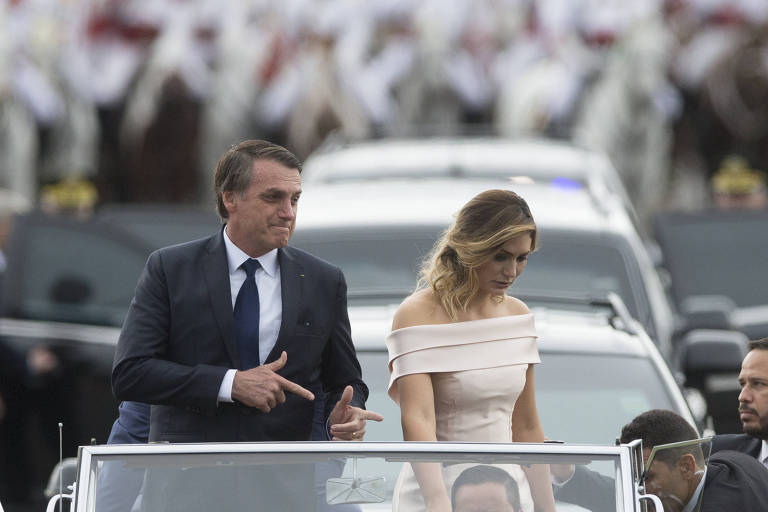 Jair Bolsonaro, de terno e gravata, faz o gesto da arma com as mãos; ao seu lado, Michelle, de vestido; eles estão em pé, em carro que os leva durante a possa