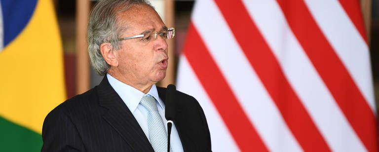 Ministro da Economia, Paulo Guedes, vestido de terno preto e gravata azul clara. Atrás dele há pedaços de bandeira do Brasil e dos Estados Unidos. Ele está falando ao microfone 
