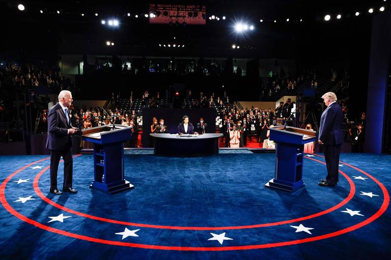 A imagem mostra dois homens em pé atrás de púlpitos em um palco circular azul com estrelas brancas e um círculo vermelho. Eles estão participando de um debate presidencial. No fundo, há uma plateia sentada e um moderador ao centro, também atrás de um púlpito.