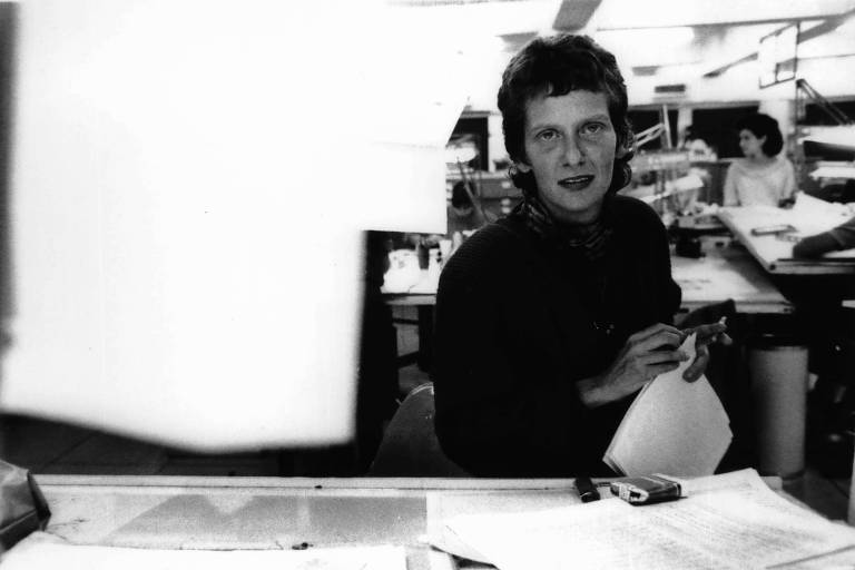 retrato em preto e branco de marilza dias costa vestindo roupas pretas e de cabelo curto, sorrindo em frente a uma mesa de trabalho 