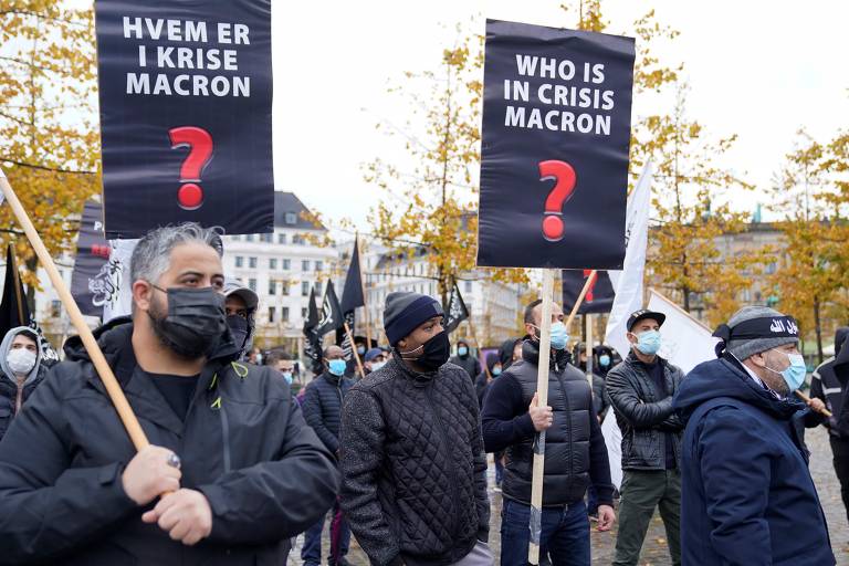 Manifestatantes seguram cartazes com a pergunta "Quem está em crise, Macron?" e símbolos do grupo muçulmano Hizb ut-Tahrir em frente a embaixada francesa na Dinamarca