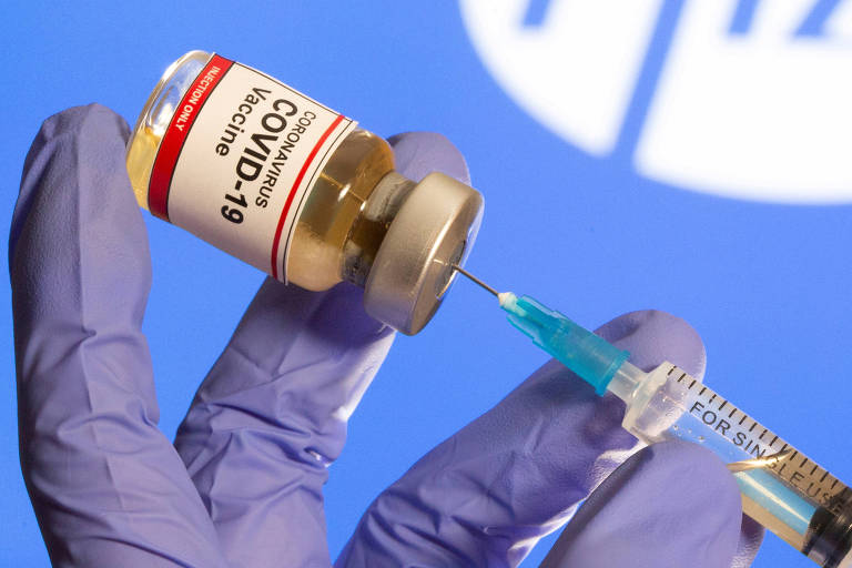 Mãos com luva azul seguram tupo de vacina no qual é espetada a agulha de uma seringa