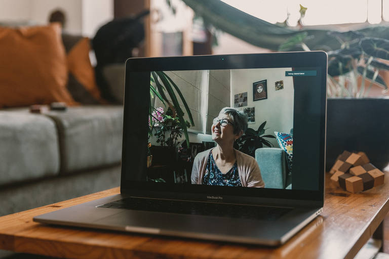 Vemos uma senhora de cabelos grisalhos curtinhos, de óculos, sorridente; ela é fotografada enquadrada na tela de um laptop, pois as imagens foram feitas a distância, a partir da casa do fotógrafo