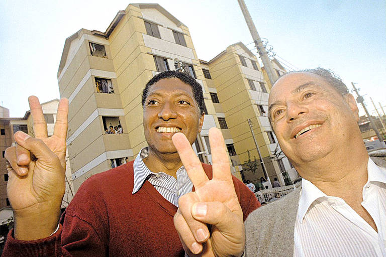 A imagem mostra dois homens sorrindo e fazendo o sinal de paz com as mãos. Ao fundo, há um edifício de apartamentos com várias janelas. O céu está claro e a iluminação é natural.