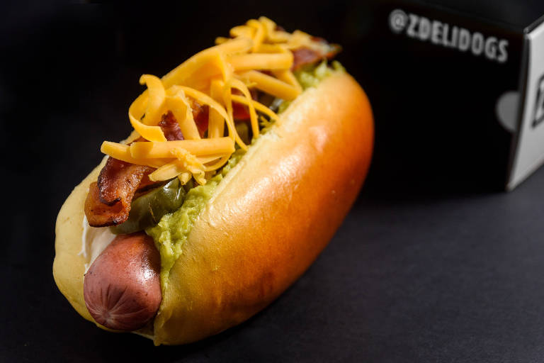 Com fundo preto, imagem mostra o hot dog com salsicha e queijo cheddar proeminentes