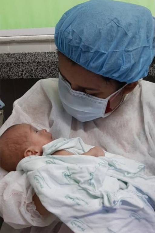 Mulher paramentada com touca, máscara e avental hospitalar segura bebê no colo