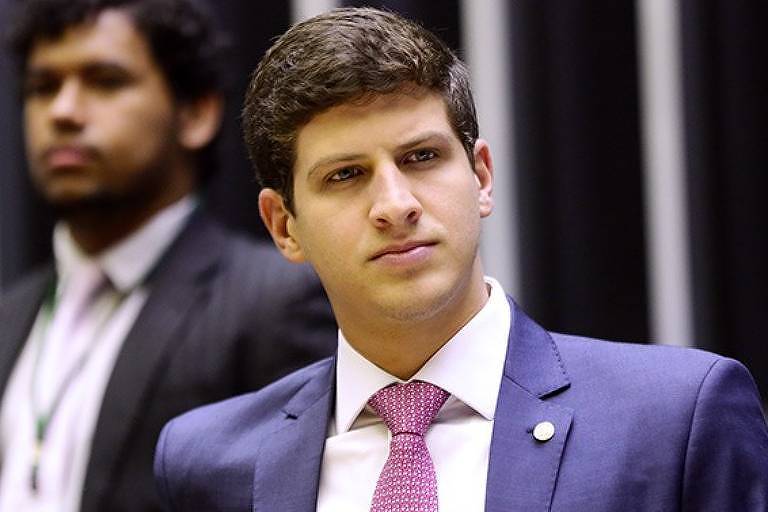 João Campos (PSB), candidato à Prefeitura do Recife, aparece em primeiro plano, de camisa branca, gravata  e paletó. Atrás, desfocado, há um outro homem de terno