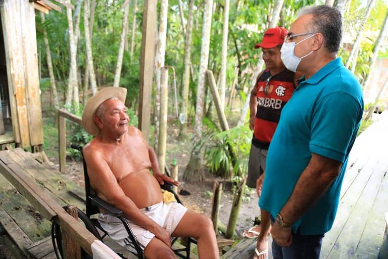 Homem de máscara e blusa azul está em frente a um idoso sem camisa, sentado, com um homem atrás com camisa do Flamengo
