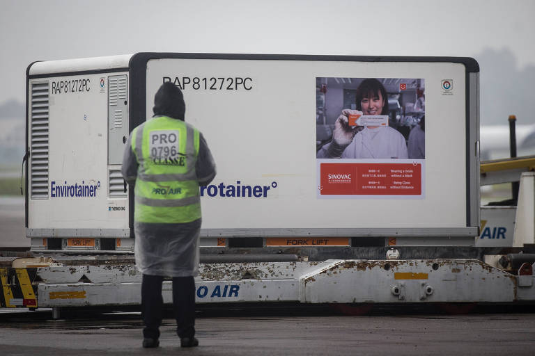 Homem de costas com colete de funcionário de pista de aeroporto observa container branco com doses de Coronavac
