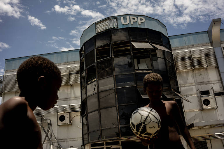 Duas crianças negras e sem camisa jogam com uma bola de futebol em frente à prédio com letreiro "UPP".