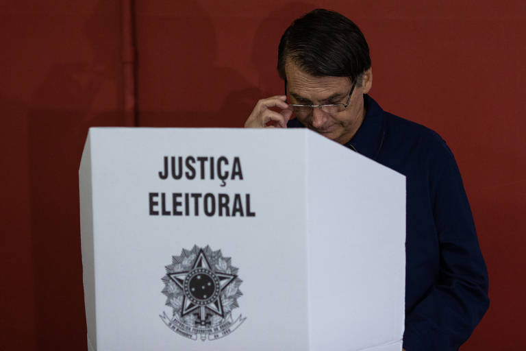 urna eleitoral com proteção onde se vê escrito Justiça Eleitoral, atrás dessa espécie de biombo, está Jair Bolsonaro,votando