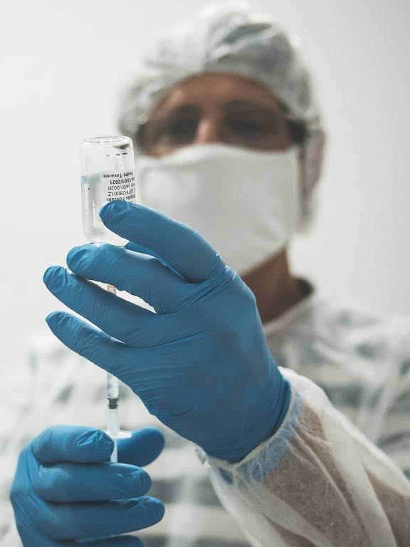 Homem com máscara e e luvas azuis retira com seringa a vacina de um pequeno recipiente