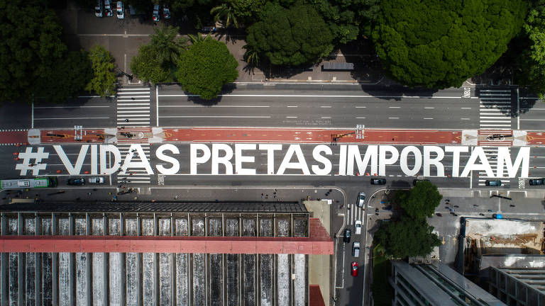Vista aérea da Avenida Paulista com a inscrição, em branco, da frase VIDAS PRETAS IMPORTAM na pista; acima aparecem árvores do Trianon; abaixo está o teto do Masp, com vigas vermelhas