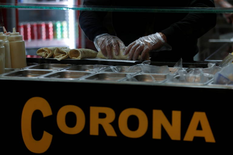 Imagem mostra as mãos de uma pessoa, com luvas, montando um sanduíche sobre um balcão onde é possível ler "corona"