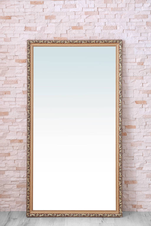 Espelho que reflete o vazio; é um espelho retangular, de corpo inteiro, com moldura dourada, toda trabalhada, e está encostado contra uma parede de tijolinhos de cor clara, quase branca; o espelho não reflete nada