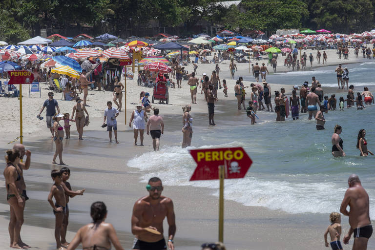 Fotografia colorida mostra uma praia lotada de banhistas. No centro da imagem há uma placa de perigo fincada na areia da praia. 