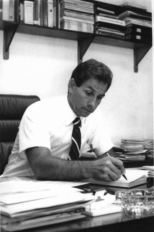 homem branco de cabelos pretos sentado em uma mesa de escritório; ele veste camisa branca e gravata e faz anotações em uma folha de papel 