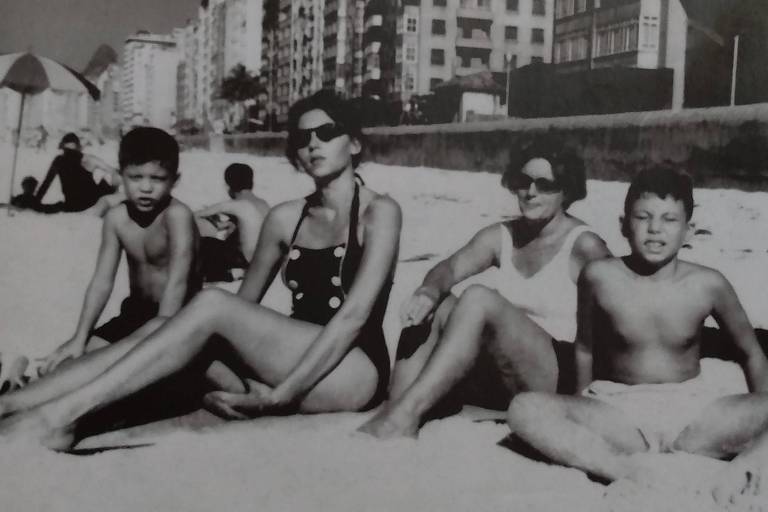 Quatro pessoas, sendo duas crianças, sentadas na areia de uma praia e olhando para a câmera