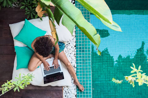 bird view of a remote online working digital nomad women with curly hair and laptop sitting crossed legged at a sunny turquoise water pool surrounded by cushions and plants in the foreground Ver menos
Foto:  shellygraphy / Adobe stock